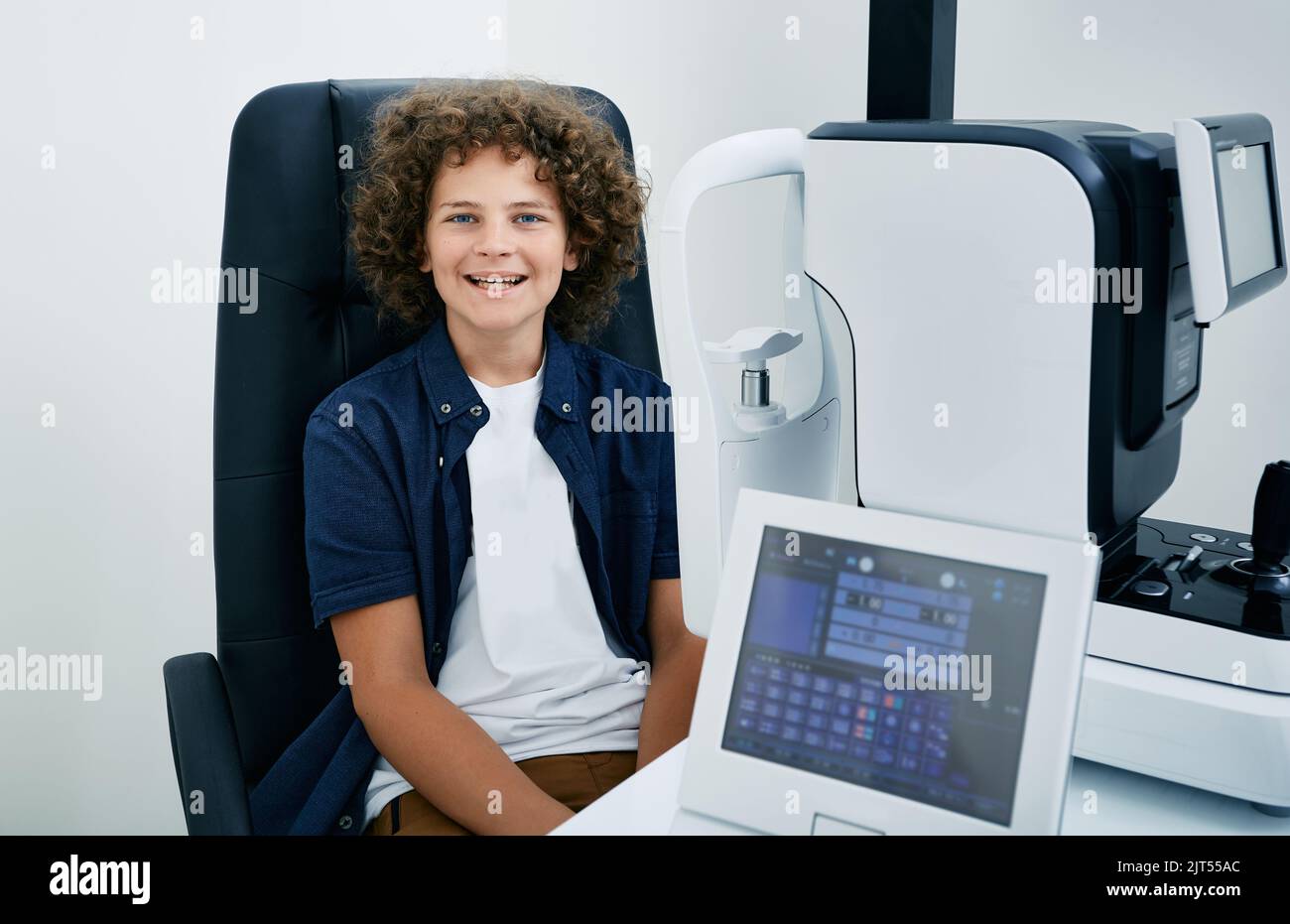 Ragazzo sorridente dai capelli ricci in ufficio oftalmologo durante la consultazione e l'esame degli occhi su moderne apparecchiature oftalmologiche Foto Stock