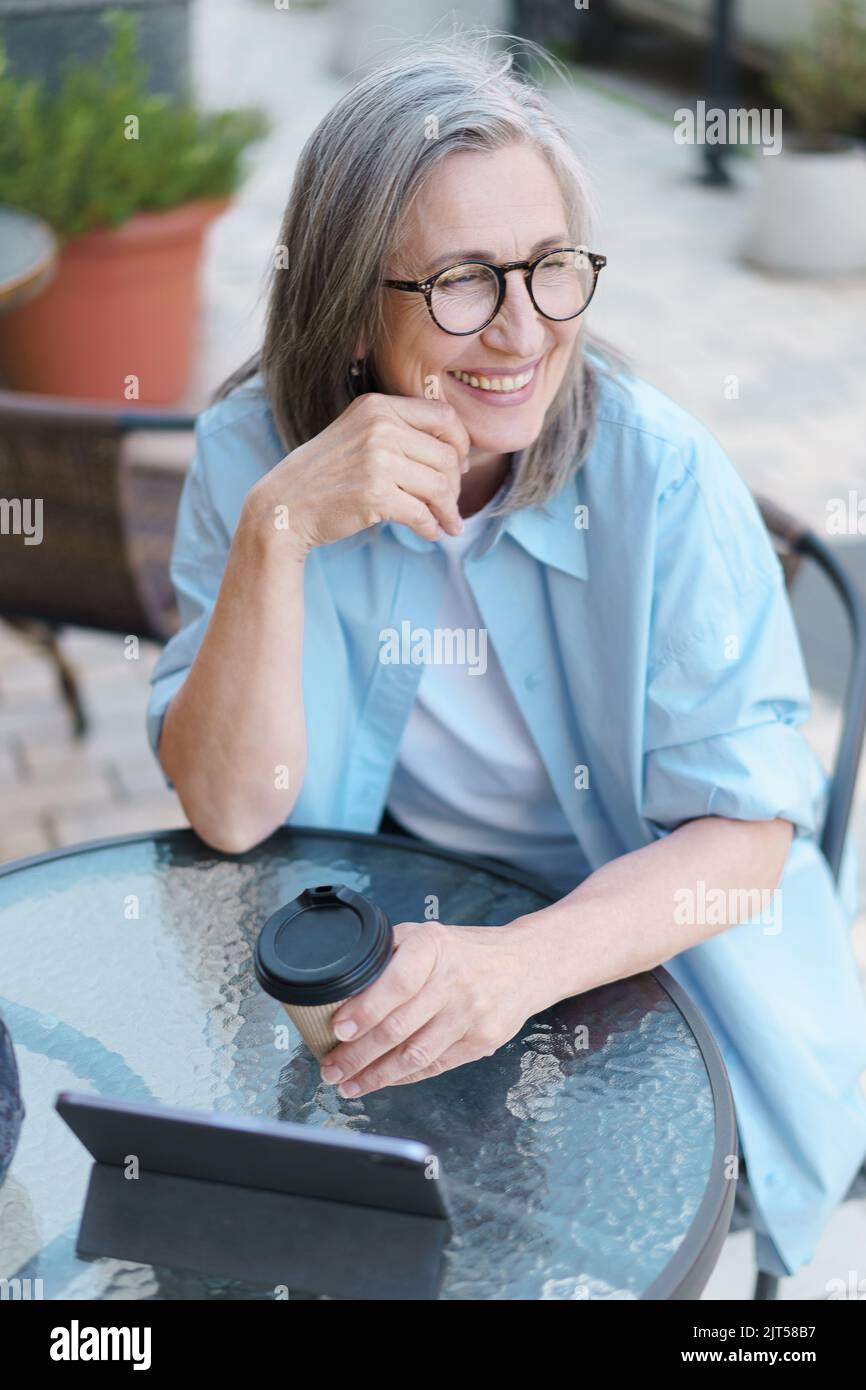 Una donna europea sorridente dai capelli grigi beve un caffè utilizzando un tablet digitale seduto al tavolo del Street cafe. Donna capigliata d'argento che lavora all'aperto City Cafe, indossando una t-shirt bianca e una camicia blu. Foto Stock