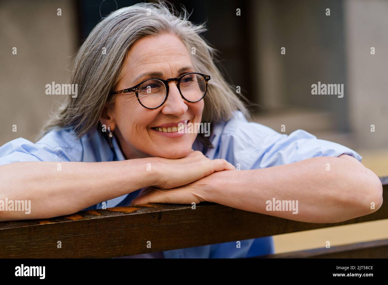 Appoggiata sulla panchina felice donna capelli grigio maturo sorridente seduta godendo il tempo estivo per le strade della città vecchia europea. La vita di pensione di svago della gente matura. Foto Stock