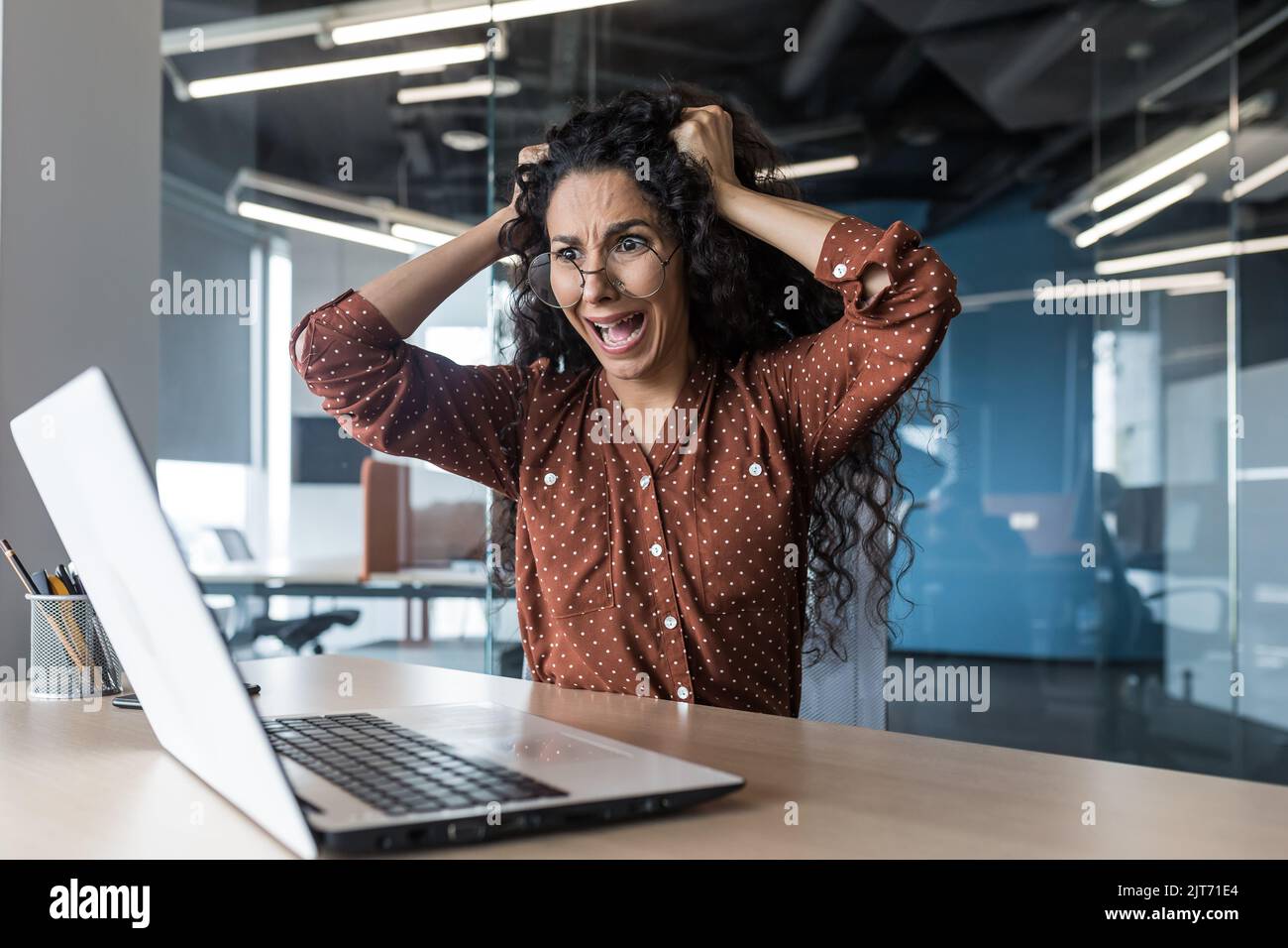 Situazione critica sul lavoro donna d'affari in shock urla e strappa i capelli sulla sua testa, delusa donna latino-americana guarda lo schermo del notebook, donna lavora all'interno dell'ufficio Foto Stock