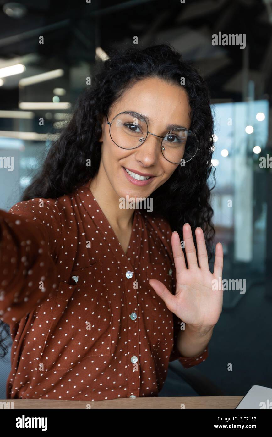 Ripresa verticale, giovane donna ispanica bella business con capelli ricci parlando in videochiamata, ondeggiando alla fotocamera dello smartphone, utilizzando l'app di comunicazione remota Foto Stock