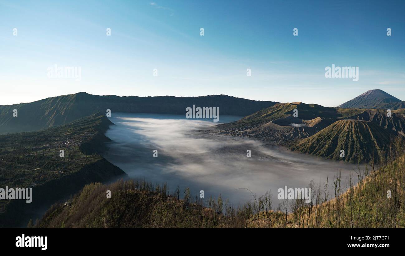 Vista panoramica mistica e surreale sulle montagne dei crateri del vulcano bromo con superficie nebbiosa e vegetazione verde, tra cui il villaggio di Cemoro Lawang durante la mor Foto Stock