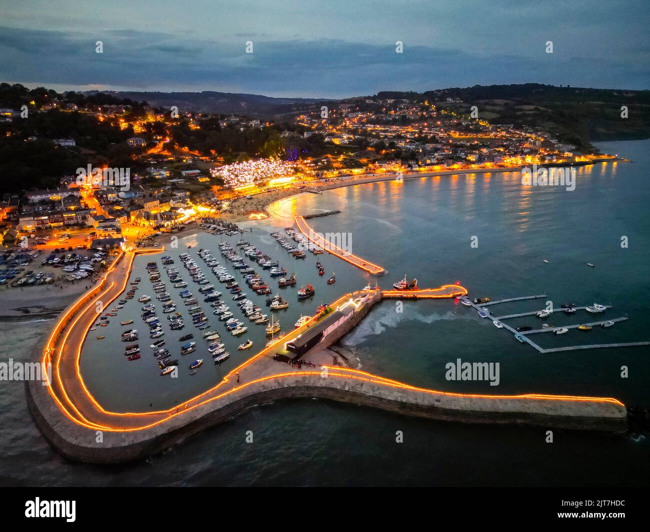 Lyme Regis, Dorset, UK. 28th agosto 2022. Vista dall'aria come migliaia di candele luce del tè illuminano le mura storiche del porto di Cobb a Lyme Regis in Dorset al crepuscolo. C'è anche una gigantesca corona illuminata sulla spiaggia per commemorare il Giubileo del platino della Regina. Questo evento spettacolare in cui la gente sponsorizza una candela in memoria di una persona amata sta raccogliendo denaro per Lyme Regis Youth Projects, British Red Cross e Cancer Research UK. Picture Credit: Graham Hunt/Alamy Live News Foto Stock