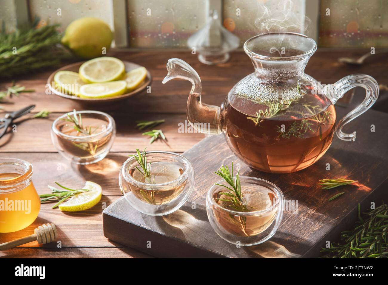 Tè al rosmarino caldo in teiera di vetro e tazze. Servito con miele e limone fresco su sfondo rustico in legno. Foto Stock