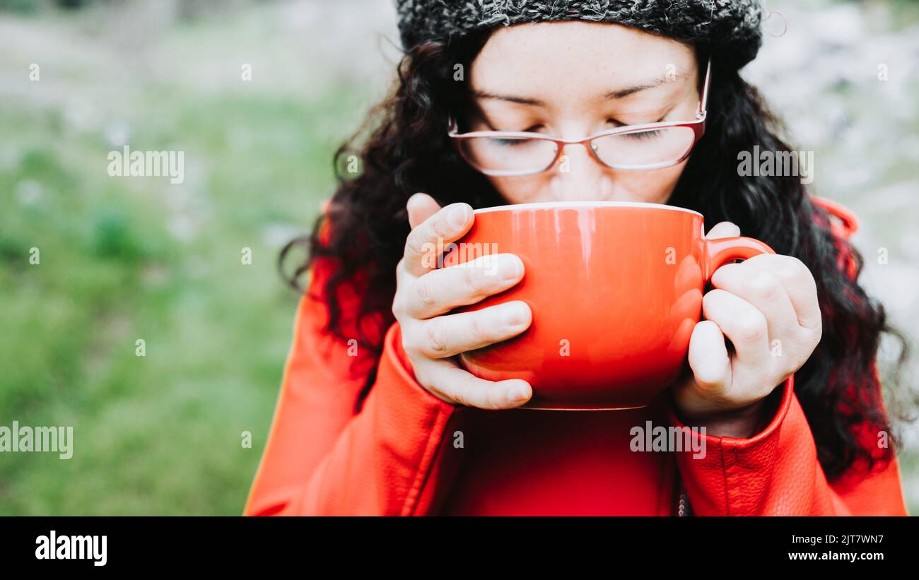 Bruna donna riccia, con giacca in pelle rossa e berretto di lana, bevendo caffè caldo in natura. Foto Stock