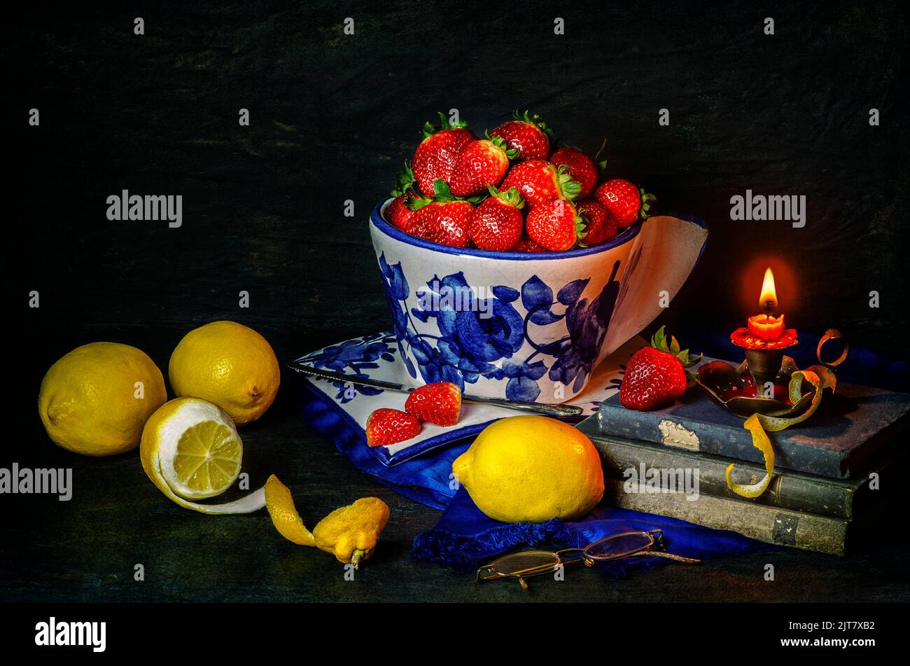 Ancora vita con deliziose fragole fresche poste in ciotola di porcellana con limoni freschi, vecchi libri e candela illuminata su sfondo scuro. Foto Stock