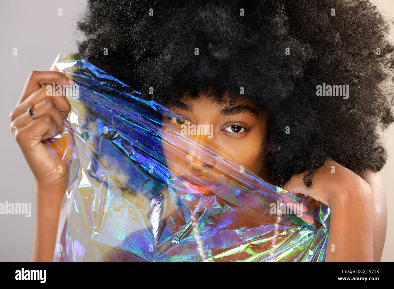 Giovane afro-americana modello femminile con capelli ricci che copre il viso con pellicola trasparente iridescente e guardando la fotocamera su sfondo grigio Foto Stock