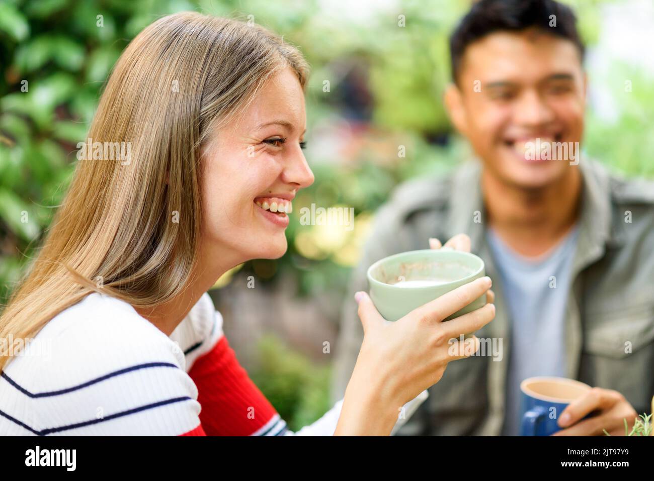Donna positiva con una tazza di cappuccino seduto sul terrazzo vicino all'uomo asiatico su sfondo sfocato durante la colazione la mattina d'estate Foto Stock