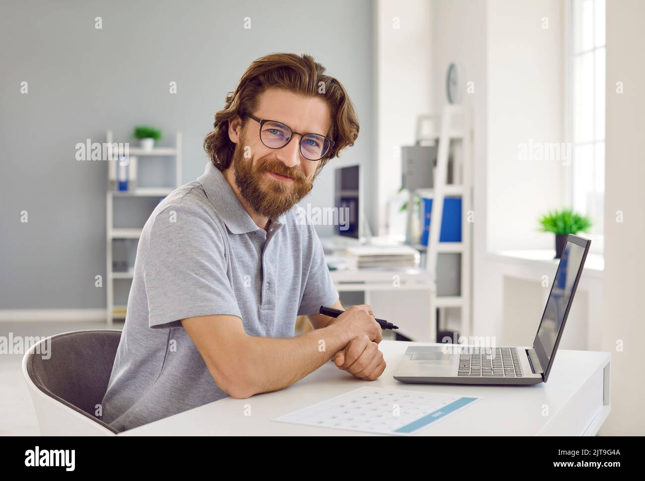Uomo felice in T-shirt seduto alla scrivania dell'ufficio con computer portatile e calendario aziendale Foto Stock