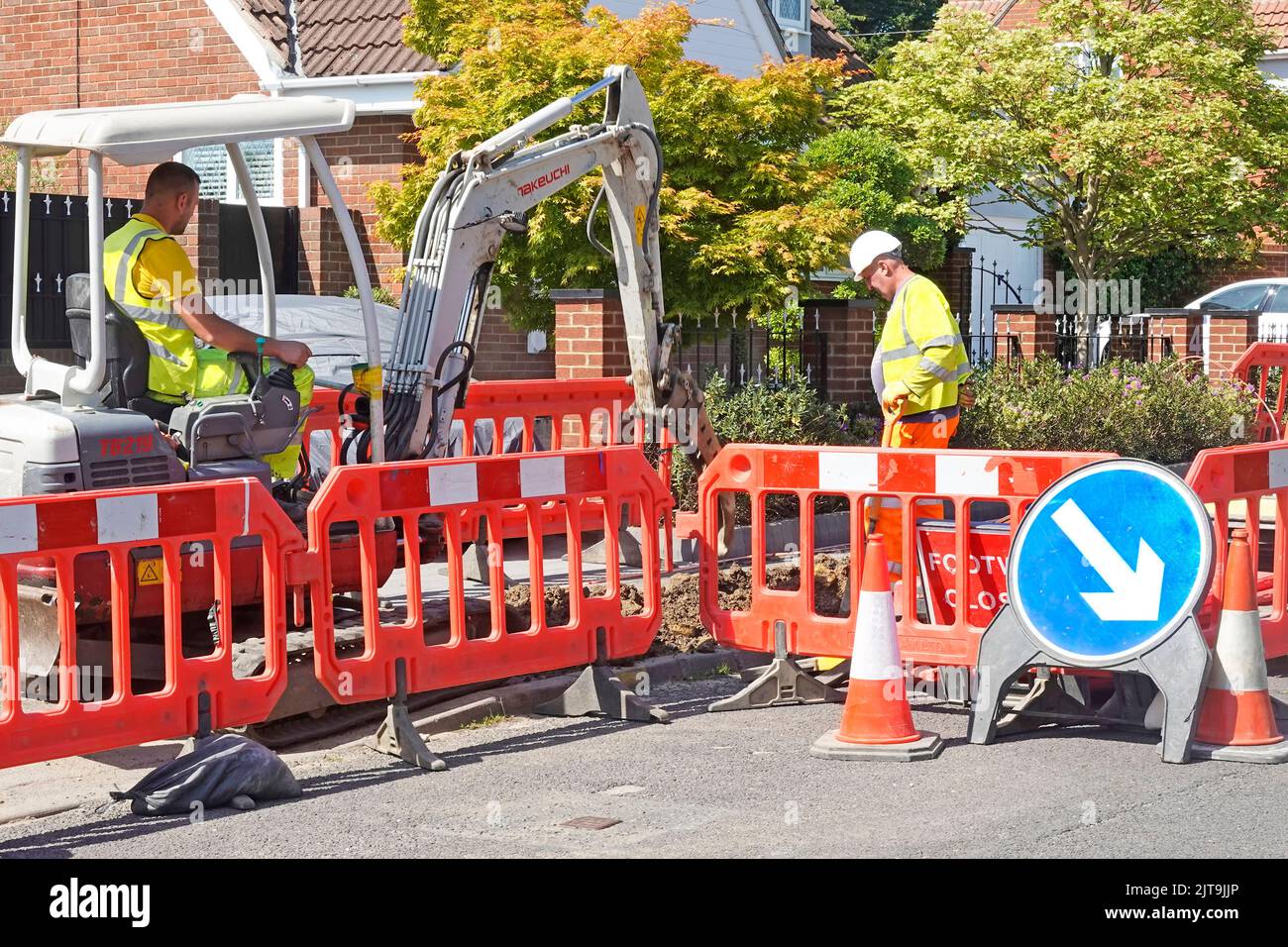 Escavatore Takeuchi mini scavatore macchina e autista uomo che lavora su strade asfaltate segnaletica barriera alta visibilità banksman in elmetto Inghilterra UK Foto Stock