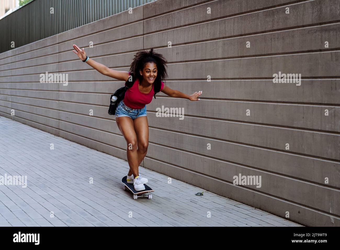 Ragazza adolescente multirazziale in sella a uno skateboard di fronte a parete di cemento, bilanciamento. Vista laterale. Foto Stock