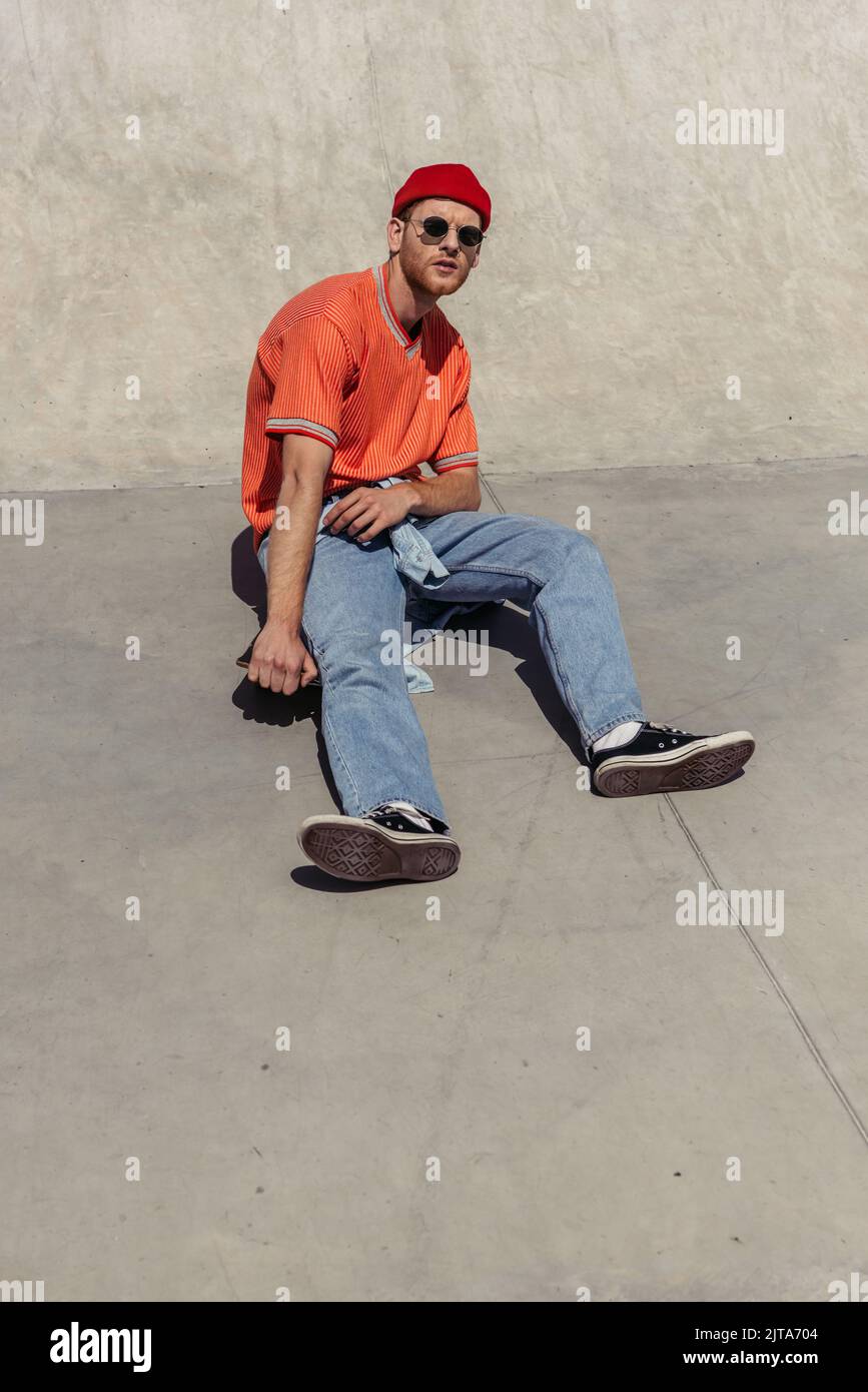 uomo a tutta lunghezza in occhiali da sole e jeans seduti su uno skate nel parco Foto Stock