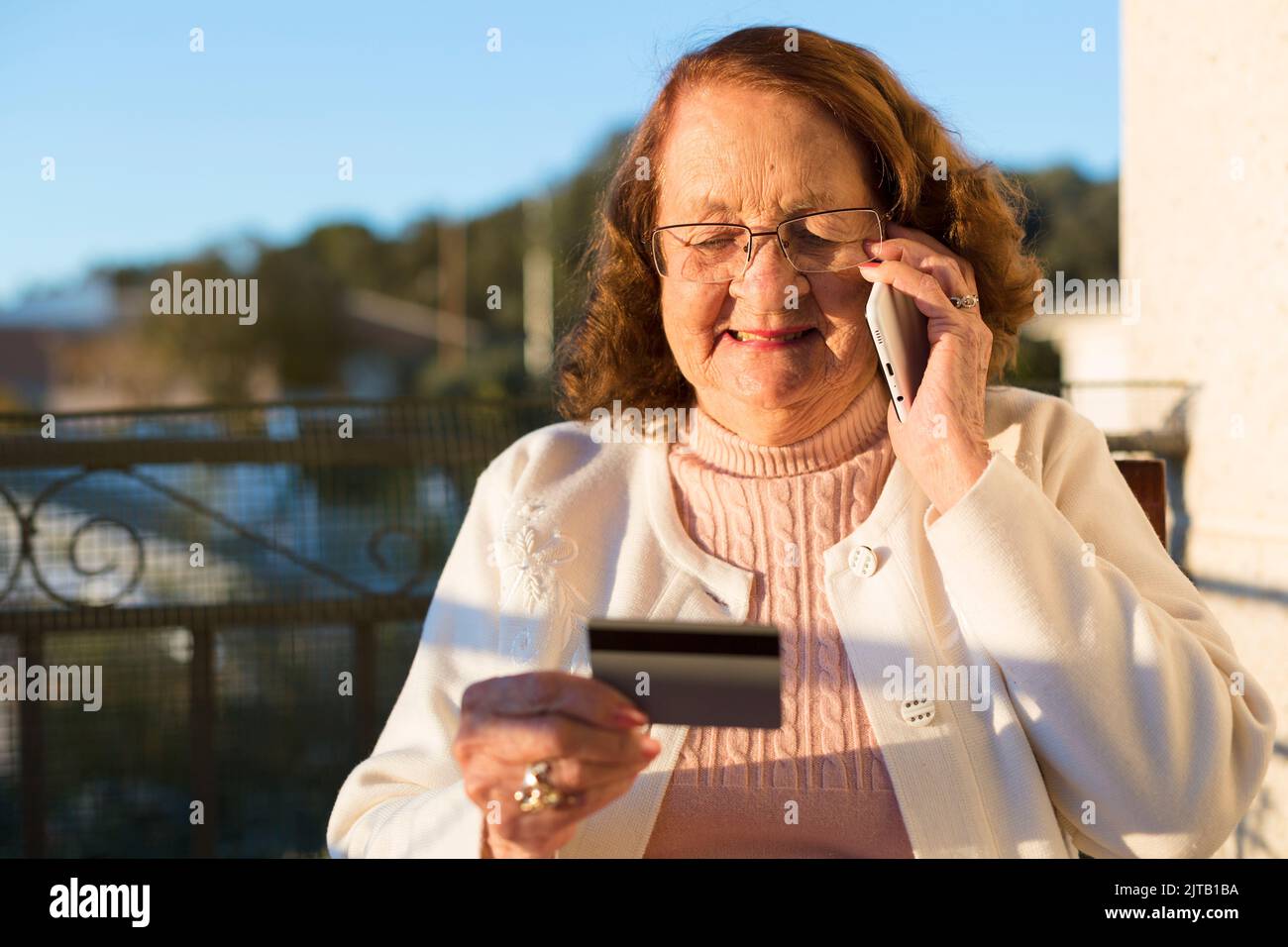 Donna caucasica anziana che effettua una chiamata telefonica tenendo in mano una carta di credito o salute. È all'aperto sulla terrazza della sua casa. Foto Stock