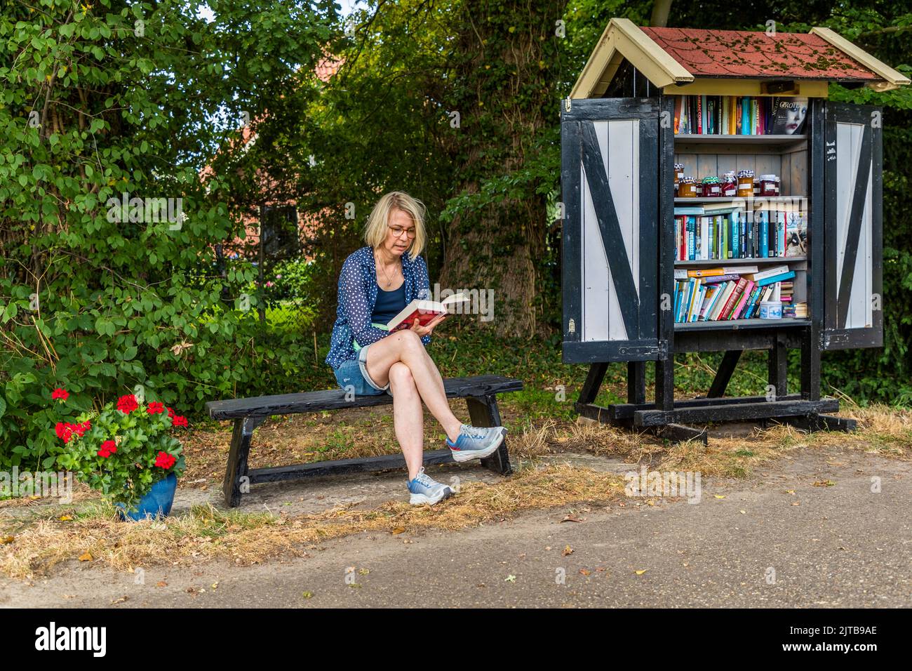 Un piccolo armadietto per lo scambio di libri sul ciglio della strada vicino ad Ambt Delden, comune di Hof van Twente, Paesi Bassi, invita i passanti a uno scambio di libri spontaneo. La giornalista Angela Berg è seduta a leggere sulla panchina accanto. Un vaso di gerani rossi è posizionato accanto alla panchina. Foto Stock