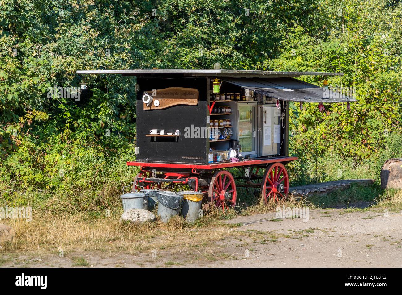 Un vecchio carrello di vendita con ruote a raggi verniciate di rosso offre marmellata, miele e prodotti caseari in un frigorifero per il self-service sul ciglio della strada vicino ad Ambt Delden, comune di Hof van Twente, Paesi Bassi. Una macchina per il caffè e contenitori per rifiuti separati per il riciclo completano l'allestimento. Foto Stock