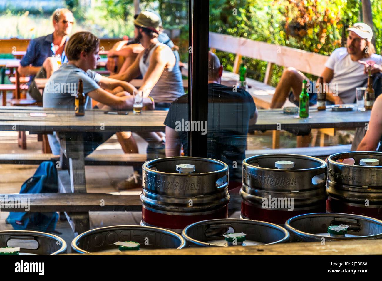 I fusti della birreria Weihenstephan si trovano in primo piano di una finestra della birreria Davo a Deventer, una delle nove città anseatiche storiche sull'IJssel, provincia di Overijssel, Paesi Bassi; dietro di loro, ospiti con bottiglie di birra sono seduti su panchine all'aperto. Foto Stock