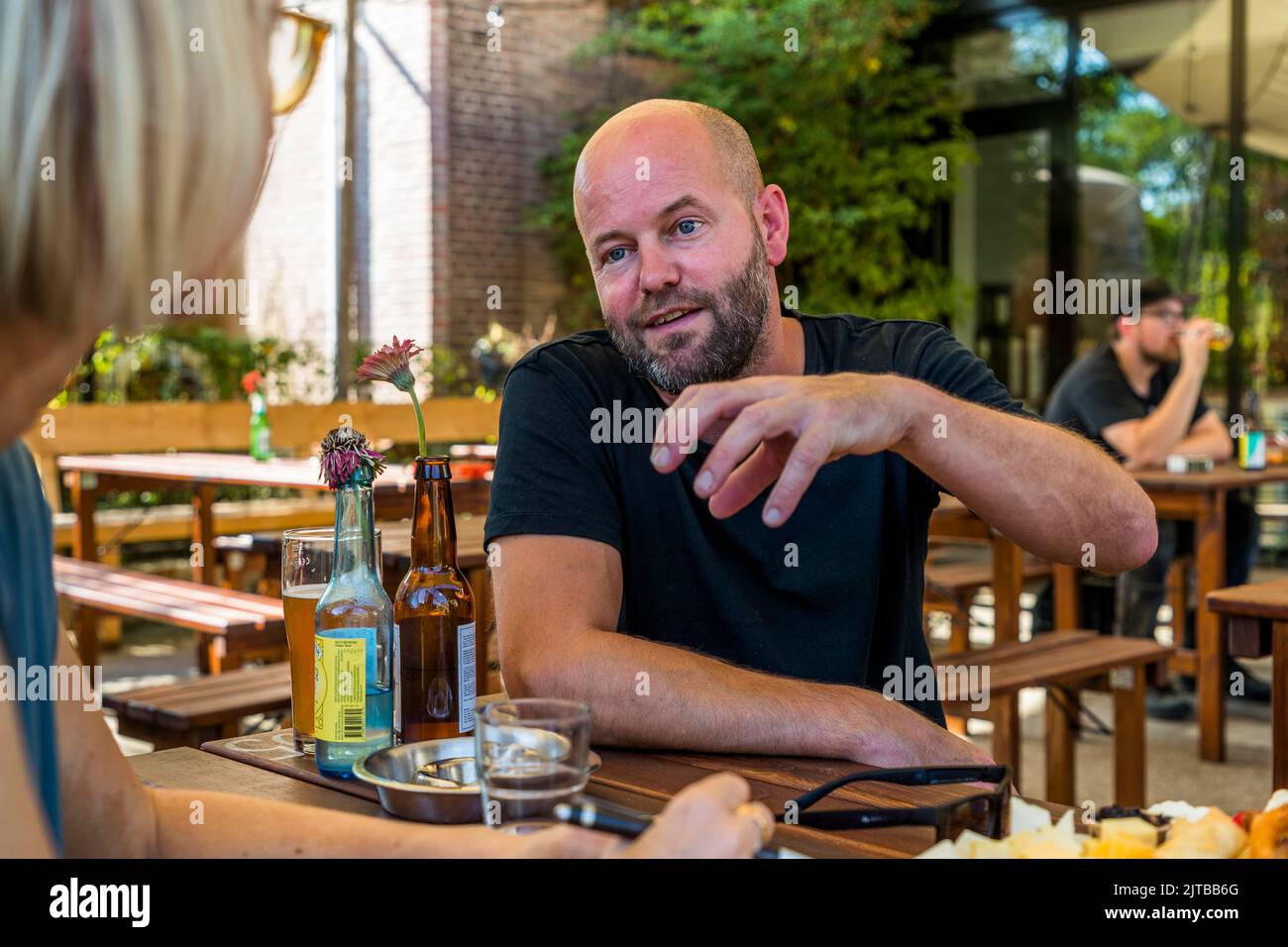 Un uomo barbuto parla alla birreria Davo di Deventer, una delle nove città anseatiche storiche sull'IJssel, provincia di Overijssel, Paesi Bassi, con la giornalista Angela Berg, di cui si vede la nuca in primo piano. Sul tavolo davanti a lui ci sono un bicchiere di birra, una bottiglia con tappo a bilico contenente un fiore e un posacenere. Foto Stock