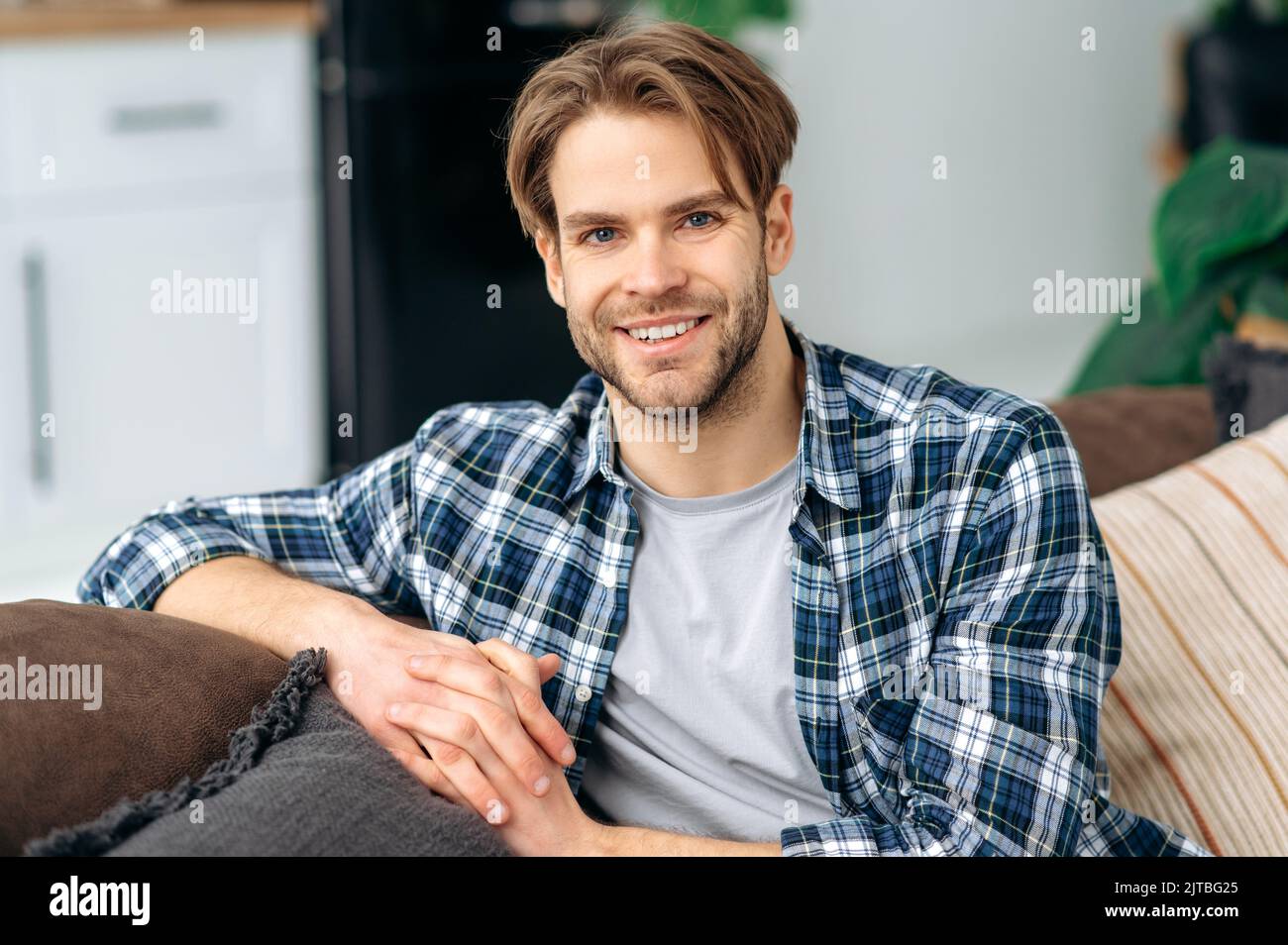 Ritratto di sicuro bel giovane caucasico dagli occhi blu, in abiti casual alla moda, seduto a casa in accogliente soggiorno sullo sfondo della cucina, guarda la macchina fotografica, sorridendo amichevole Foto Stock