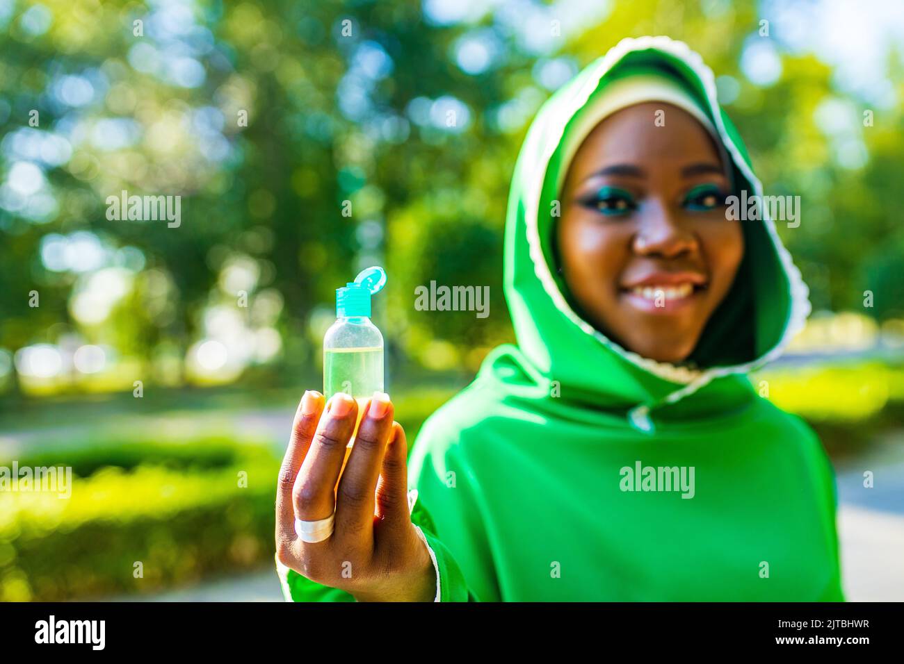 razza multiculturale donna musulmana in hijab verde e naso piersing applicare un gel antisettico all'aperto nel parco estivo Foto Stock