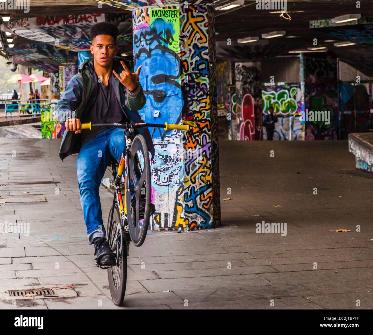 Un giovane nero che guida la sua moto su una ruota fa gesto con le dita verso la macchina fotografica all'Undercroft Skate Park in South Bank, Londra, Inghilterra, Regno Unito Foto Stock