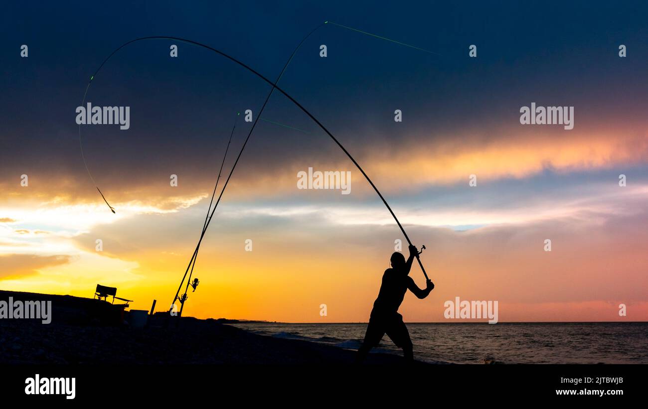 Silhouette di un pescatore che getta una canna sulla costa al tramonto. Pesca notturna con allarmi illuminati. Foto Stock