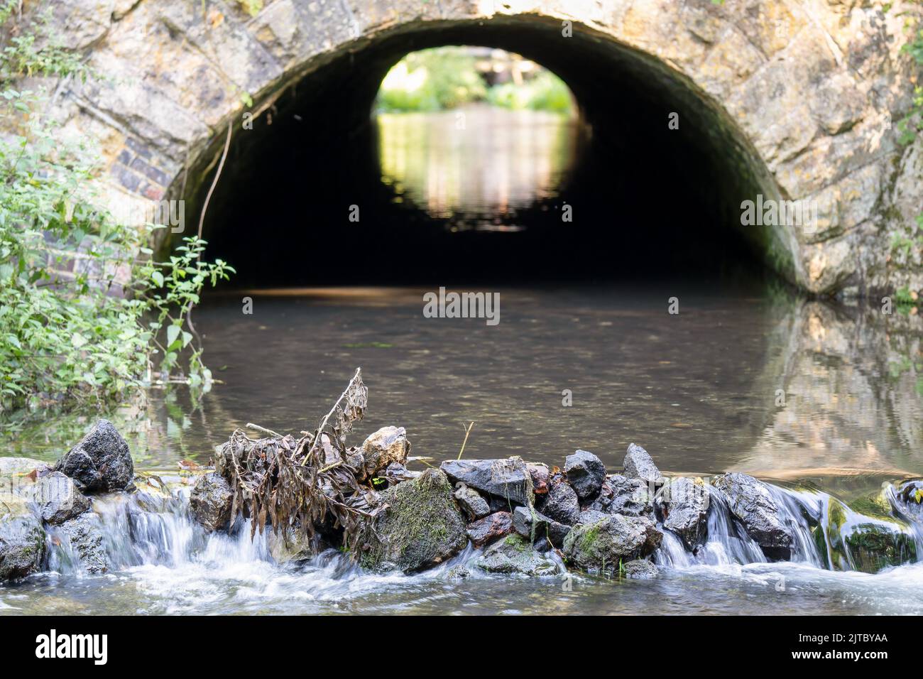 primo piano di un torrente di gesso che scorre fuori da un tunnel ad arco di pietra e sopra una piccola cascata di pietra Foto Stock