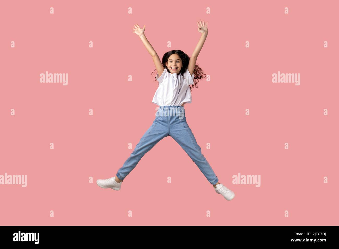 Ritratto a tutta lunghezza di bella bambina affascinante vestendo una T-shirt bianca saltando in aria e braccia sollevate, esprimendo l'emozione, divertendosi. Studio in interni isolato su sfondo rosa. Foto Stock