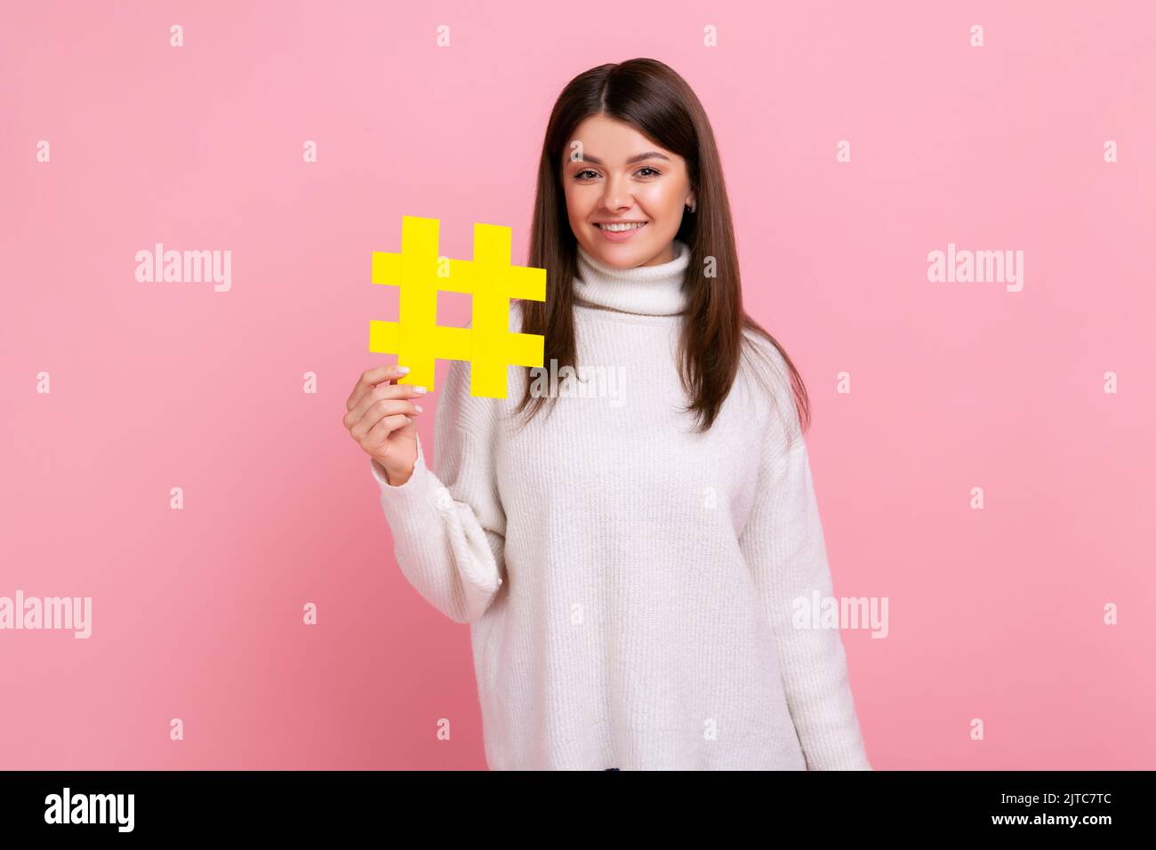 Bella donna in possesso di hashtag simbolo, promuovendo l'argomento virale in rete sociale, tagging blog tendenze, indossando bianco maglione casual stile. Studio in interni isolato su sfondo rosa. Foto Stock
