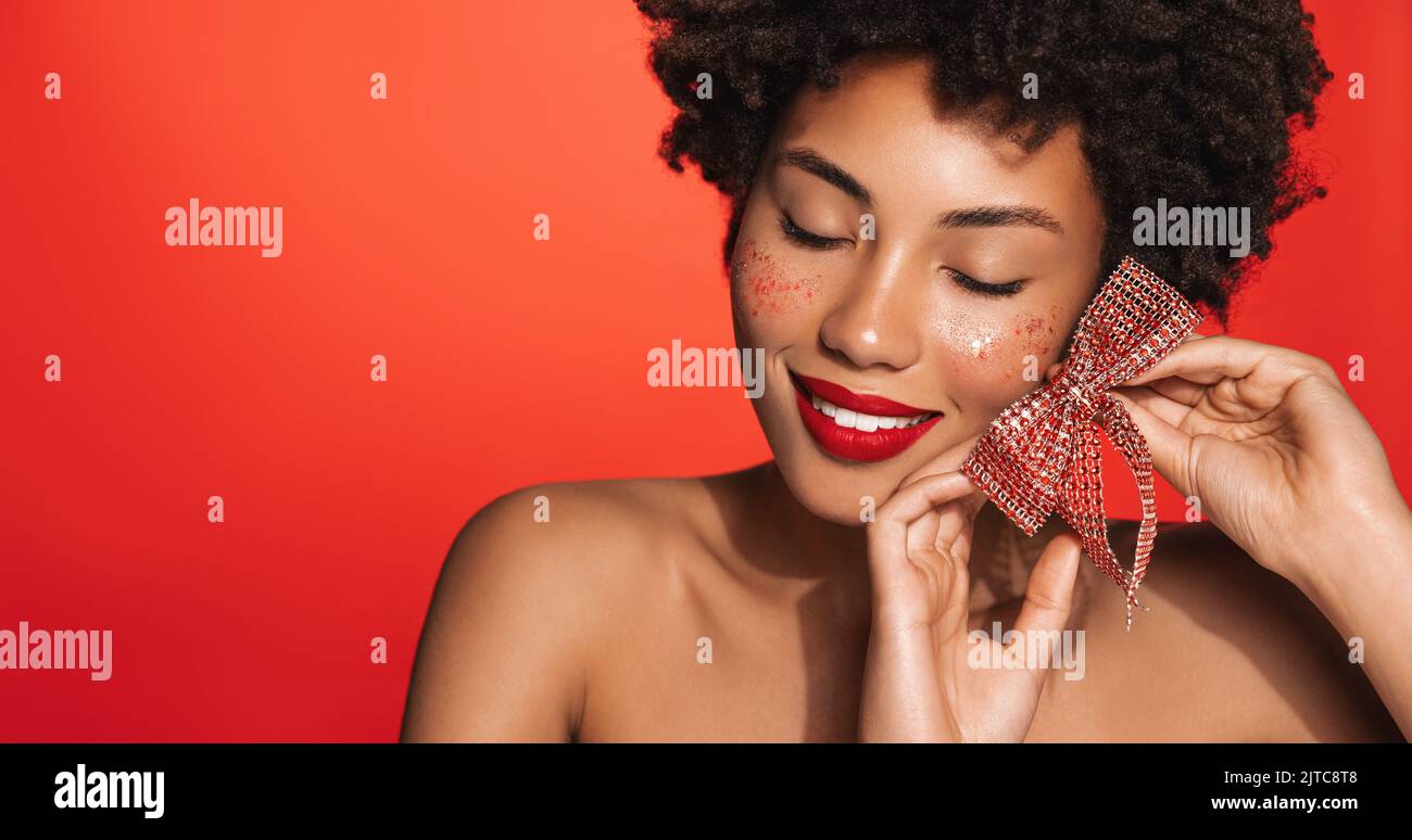 Ritratto di sorridente nero modello femminile con glitter, guardando verso il basso sensuale, mostrando bowtie, posando su sfondo rosso Foto Stock