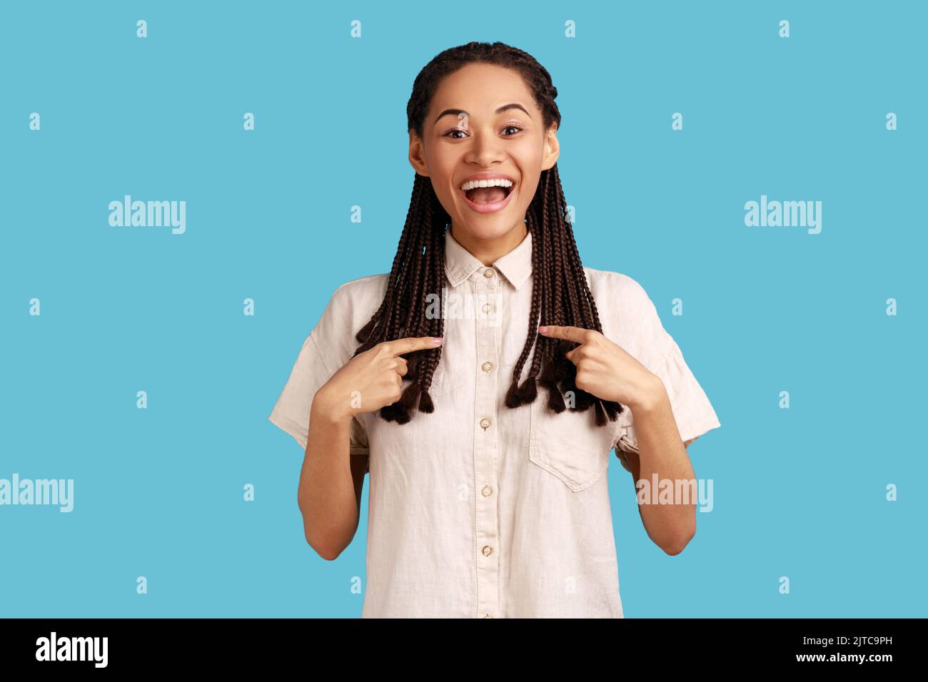 Donna allegra eccitata con greggi neri che puntano a se stessa e sorridendo in generale, estremamente felice di essere scelto, indossando una camicia bianca. Studio in interni isolato su sfondo blu. Foto Stock