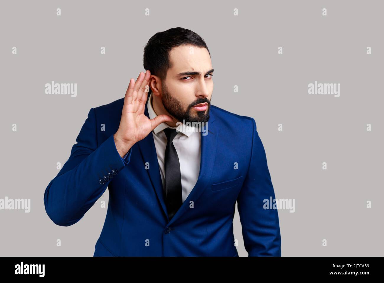Uomo bearded concentrato nosy che tiene la mano vicino all'orecchio, ascoltando i discorsi interessanti e segreti privati, spionaggio, indossando il vestito ufficiale di stile. Studio in interni isolato su sfondo grigio. Foto Stock