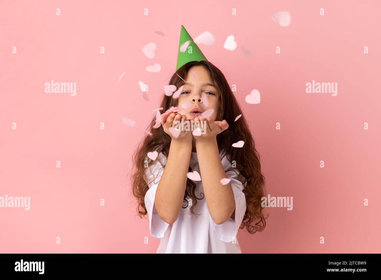 Ritratto della bambina che indossa una T-shirt bianca e un cappello da festa a forma di cuore scintillante, che si gode il compleanno, festeggia l'evento, l'umore festivo. Studio in interni isolato su sfondo rosa. Foto Stock