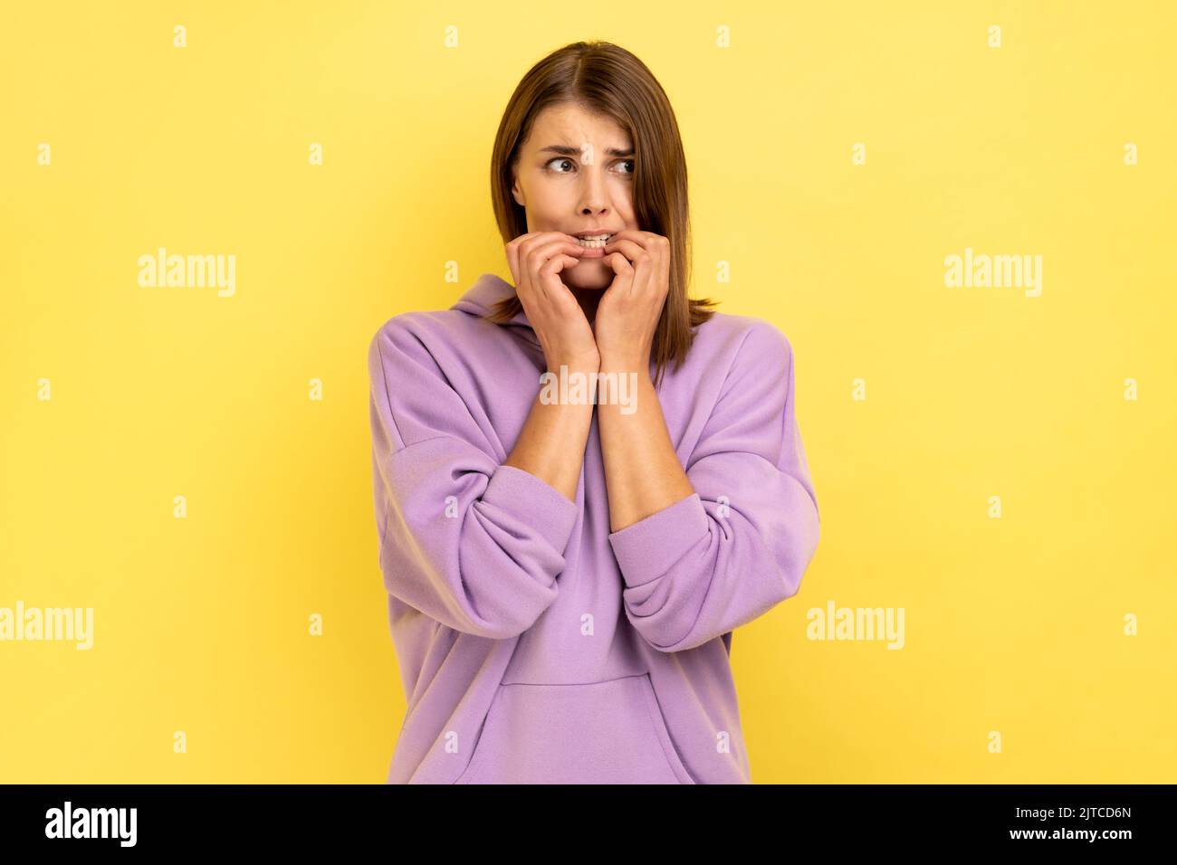 Problemi e preoccupazioni. Ritratto di donna nervosa mordente unghie, terrorizzato per problemi, fobia sofferenza, disturbo d'ansia, con felpa con cappuccio viola. Studio al coperto isolato su sfondo giallo. Foto Stock