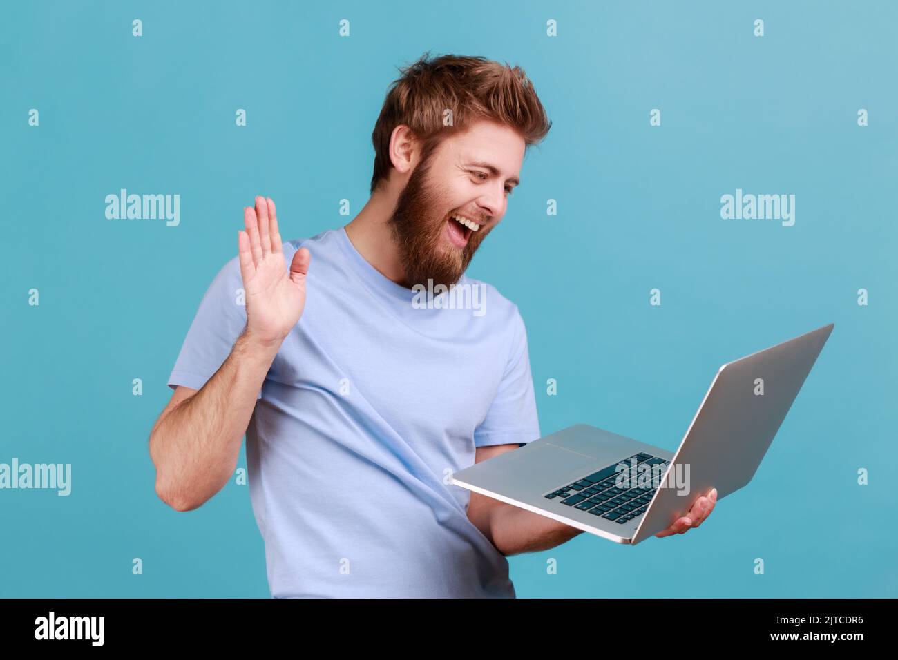 Ritratto di un uomo amichevole barbuto che saluta un lavoratore attraverso una webcam e mostra palmo del gesto della mano con un sorriso toothy, avendo conversazione in linea. Studio in interni isolato su sfondo blu. Foto Stock