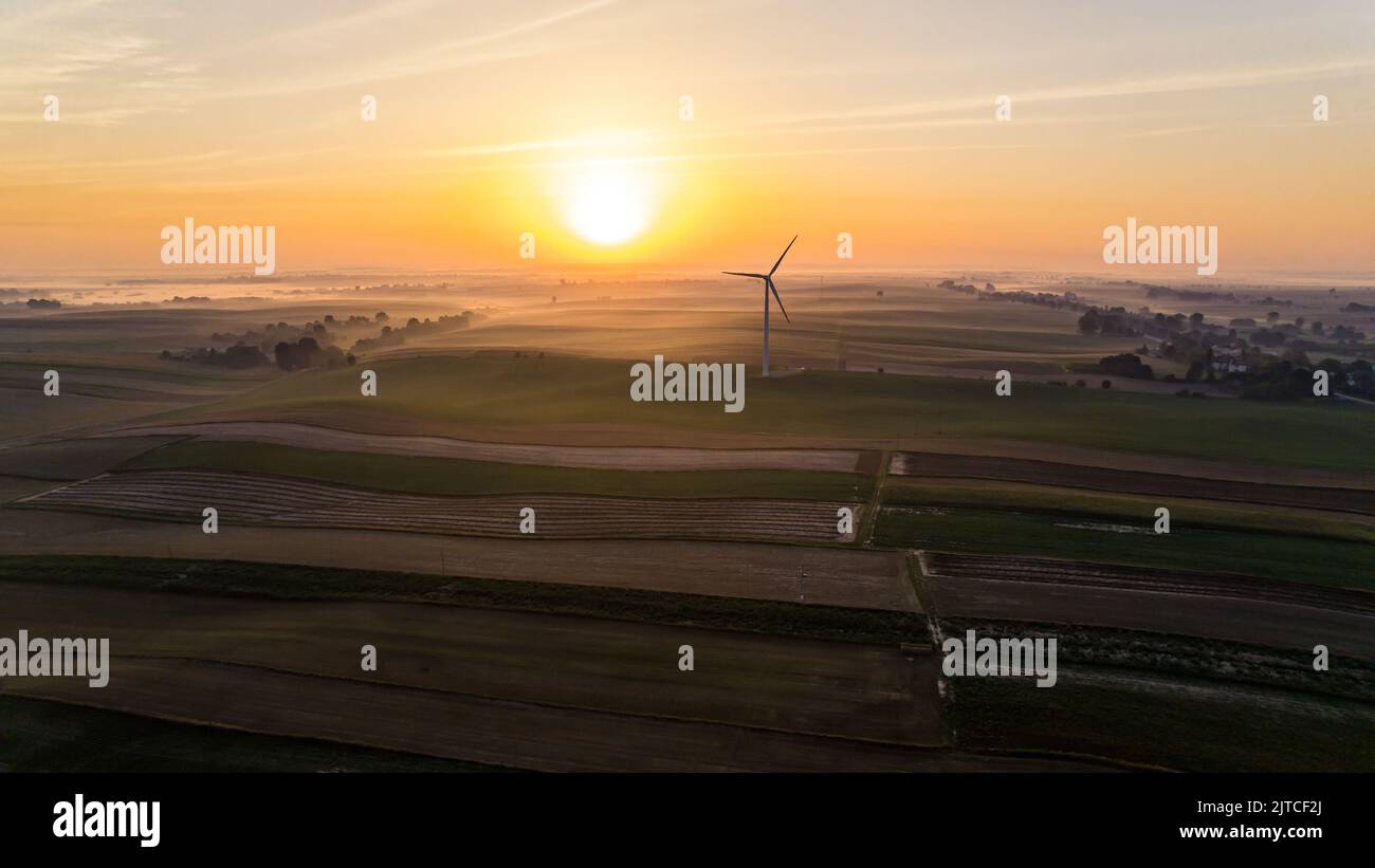 Alba colorata su campi agricoli panoramici con orizzonte nebbioso e enorme turbina eolica. Luce solare brillante. Consapevolezza ambientale. Scatto orizzontale. Foto di alta qualità Foto Stock