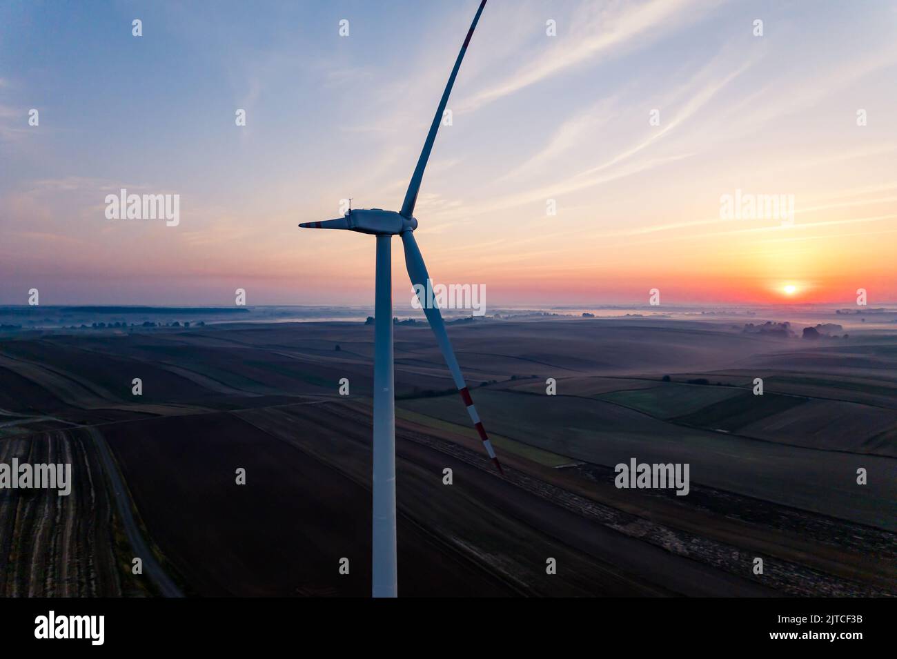 Enorme trubina del vento con campi sullo sfondo e colorato cielo all'alba con sfondo nebbia. Tecnologia moderna e rispettosa dell'ambiente. Scatto orizzontale. Foto di alta qualità Foto Stock