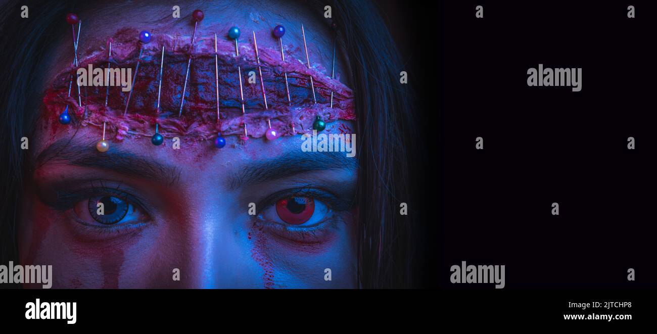 primo piano di una giovane donna con trucco horror per halloween con una grande ferita e pin sulla sua fronte e diversi occhi colorati Foto Stock