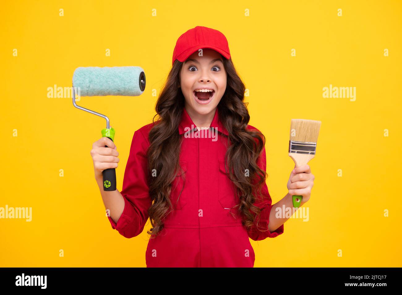 Ragazza teenager costruttore eccitato nella costruzione di uniforme e tappo. Pittore con pennello o rullo per pittura. Lavoratore isolato su sfondo giallo Foto Stock