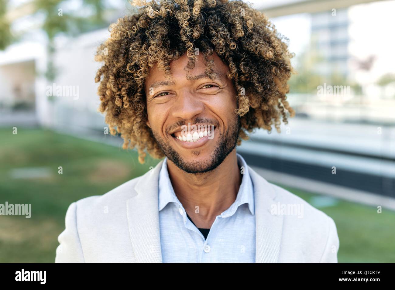 Primo piano ritratto di un felice carismatico sicuro capelli ricci brasiliano o ispano maschio, imprenditore di successo, uomo d'affari, in piedi all'aperto, guardando la fotocamera, sorridente amichevole Foto Stock