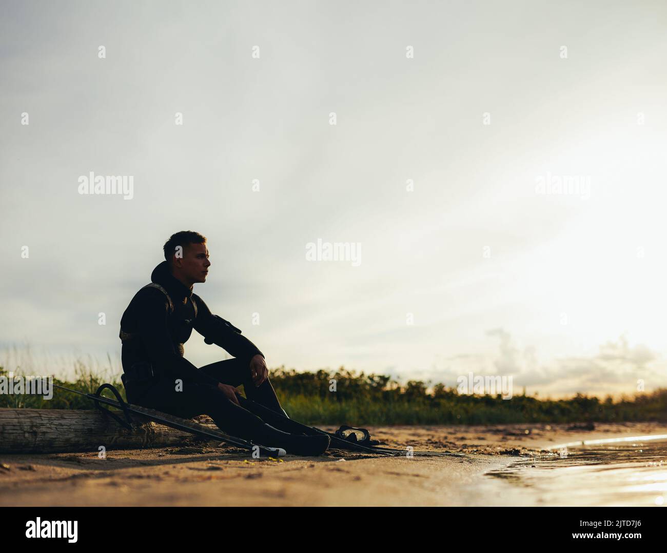 Giovane uomo avventuroso seduto sulla spiaggia in una muta. Tuffatore che guarda il mare prima di andare a pesca di spearfishing al tramonto. Foto Stock