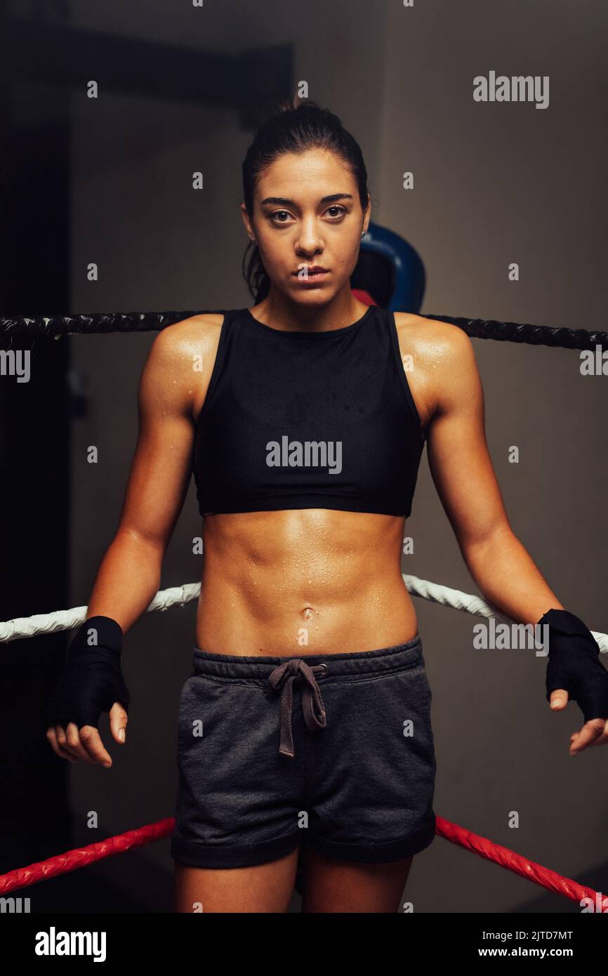 Atleta femminile che guarda la fotocamera mentre si trova in un angolo di un anello di pugilato. Pugile femminile fiducioso che si prepara per una partita di boxe. Foto Stock