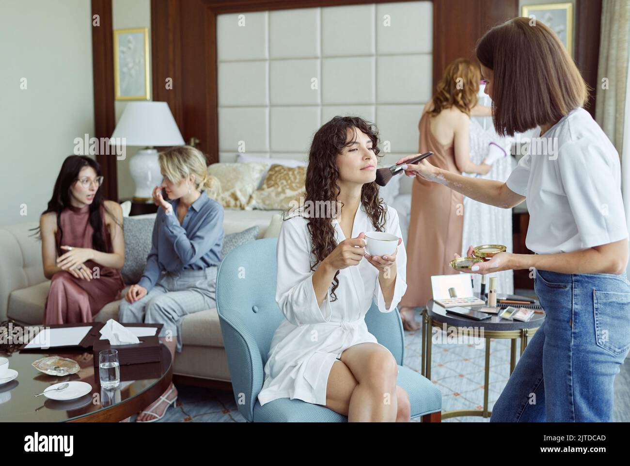 Giovane donna splendida brunetta in accappatoio bianco che ha una tazza di tè mentre si siede in poltrona di fronte al makeup artista Foto Stock