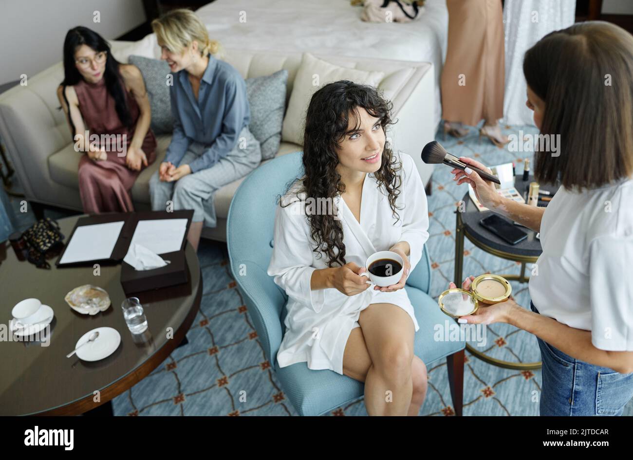 Artista visage femminile con polvere compatta e pennello applicare il trucco sul viso della giovane sposa con tazza di americano il mattino del matrimonio Foto Stock