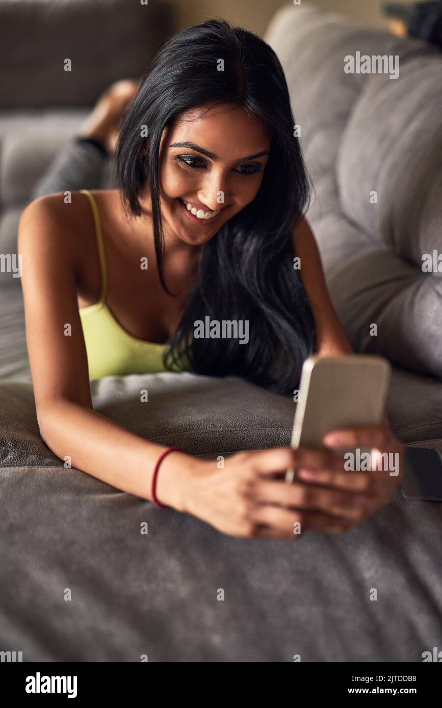 SMS nella chat di gruppo. Una giovane donna attraente che invia un SMS mentre si sdraia sul divano a casa. Foto Stock