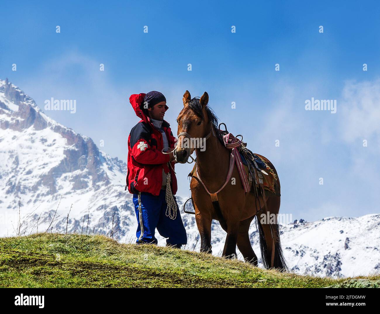 Kazbegi, Georgia - 02 maggio 2017: Un cavallo non identificato si prende cura del suo cavallo sadled sullo sfondo di vette innevate Foto Stock