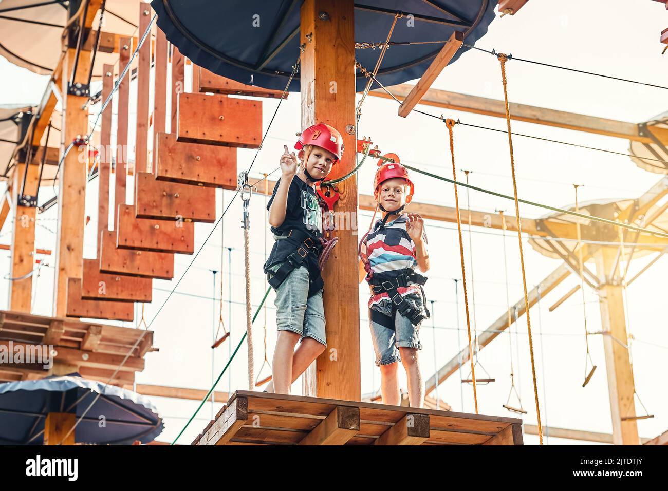 Fratelli scolari in un casco e attrezzatura per arrampicata in piedi su una piattaforma di legno contro il cielo, trascorrere il tempo nel parco di corda nella sera d'estate Foto Stock