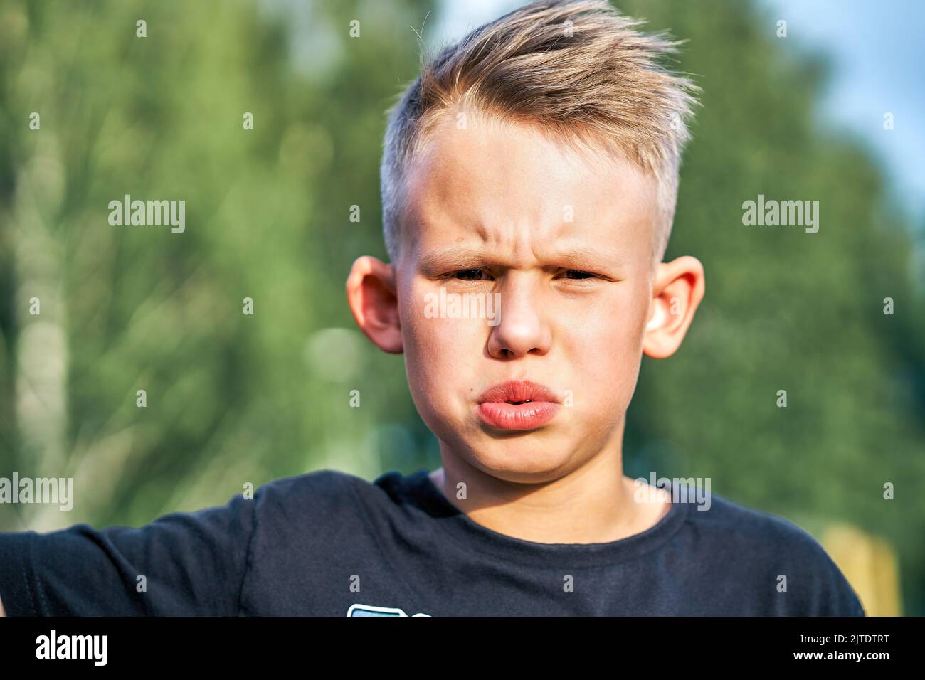 Bambino frustrato con una breve acconciatura in posa per la fotocamera verde offuscato soleggiato estate parco cittadino estrema closeup Foto Stock