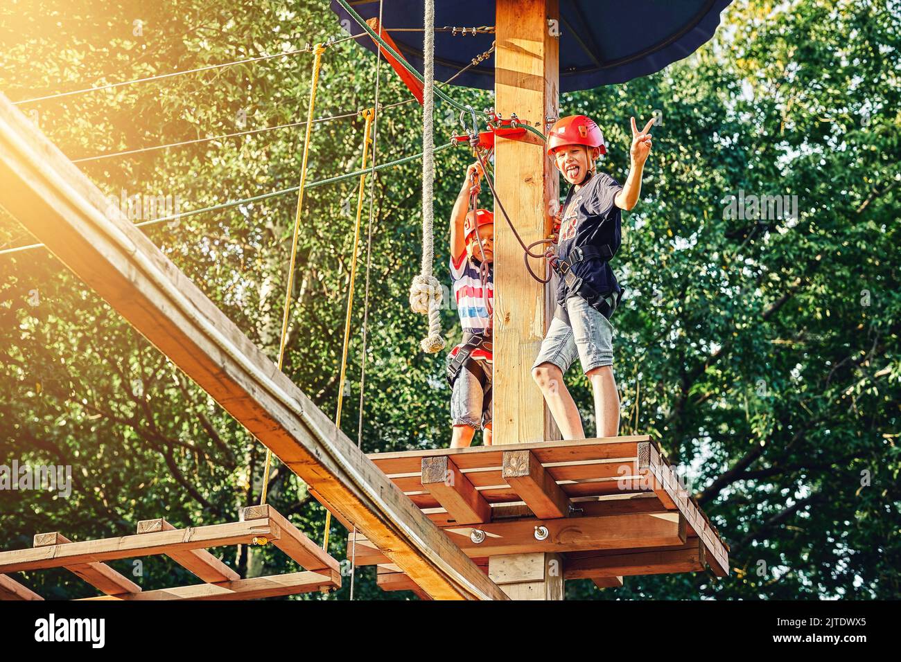 I bambini giovani dei fratelli trascorrono del tempo nel parco delle corde. Il fratello minore rafforza il moschettone di sicurezza. Il fratello maggiore mostra lingua e V gesto Foto Stock