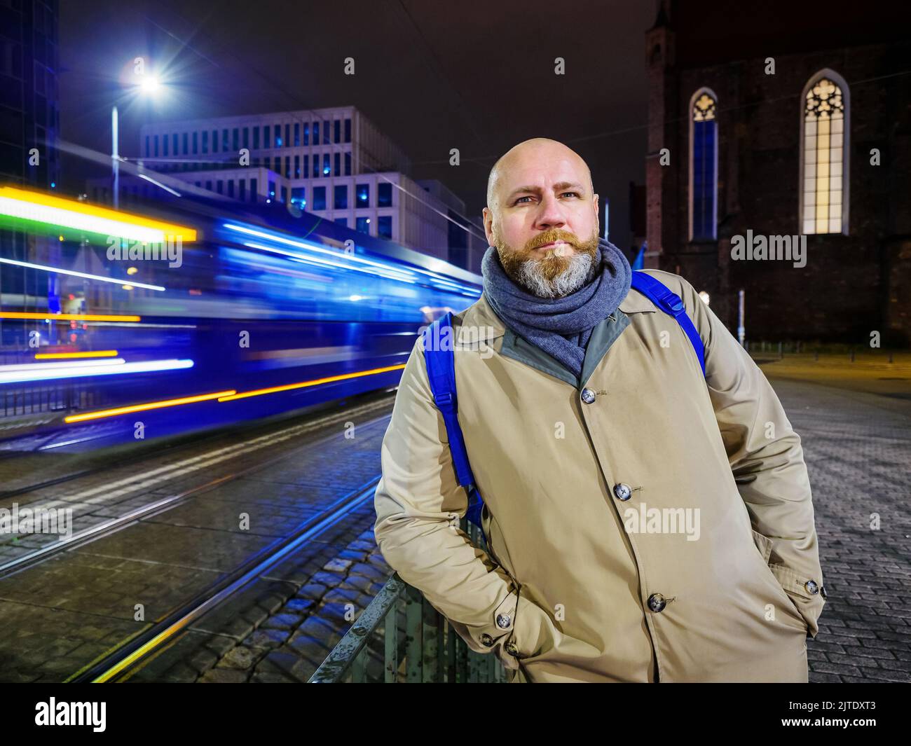 una persona adulta è in piedi da sola presso una fermata del tram in una città notturna e il tram si muove sullo sfondo Foto Stock