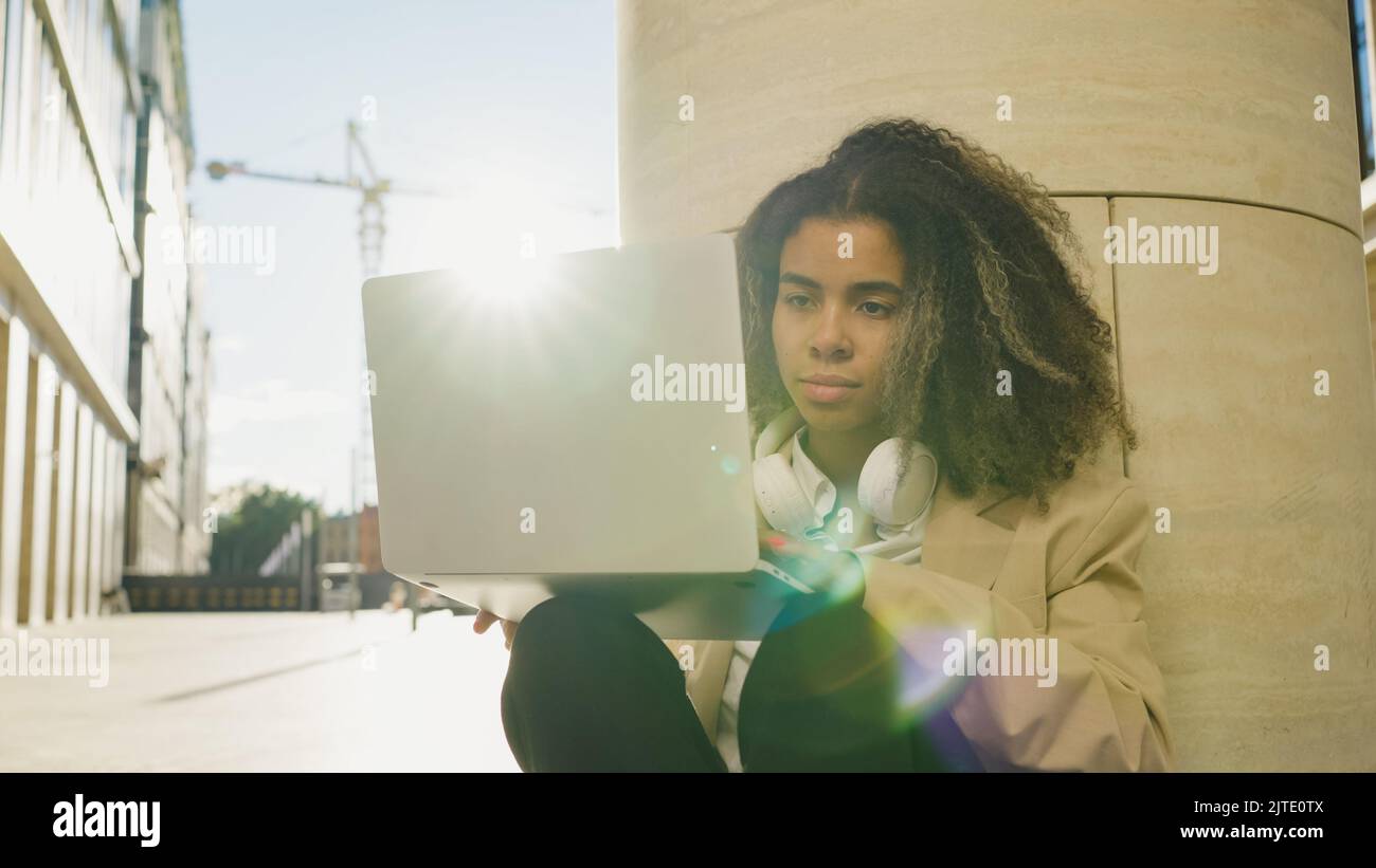 Il dipendente afro-americano inserisce le informazioni nel computer portatile seduto in strada in una luce posteriore. La giovane donna trova ispirazione al di fuori del caos dell'ufficio appoggiandosi sulla colonna Foto Stock
